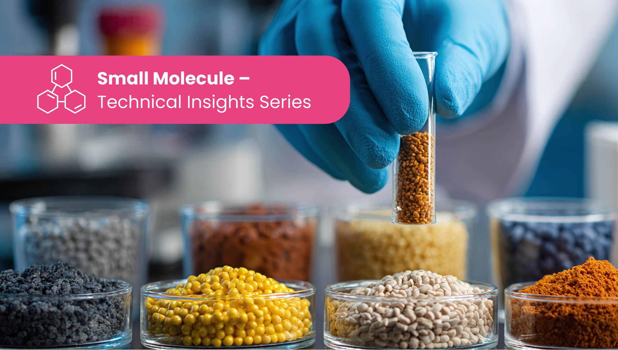 Blue-gloved hand holding a small glass vial of tan granules above an array of petri dishes filled with multicolored powders and pellets in a laboratory setting.